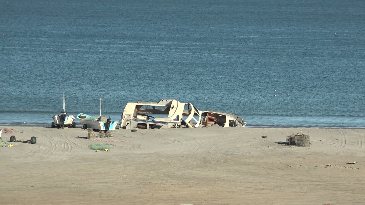 At 0:02 in San Juanico, the damaged white hull sits close to the waterline with small boats and scattered objects spread across the sand, backed by calm blue ocean and no visible people.