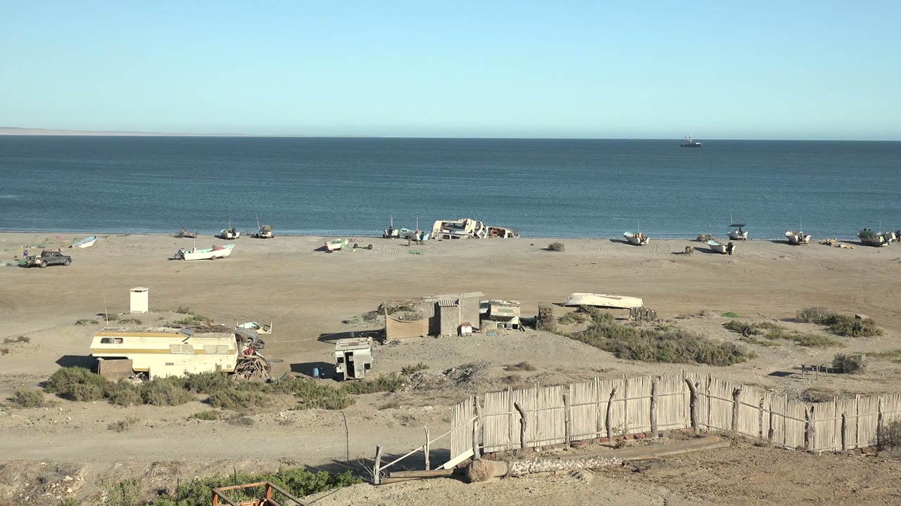 Wide ground-camera view of San Juanico, Baja California Sur with abandoned boats on a sandy beach, calm ocean beyond, a weathered fence in front, and no visible people.