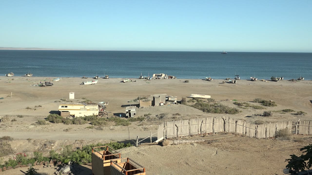 Frame from San Juanico, Baja California Sur showing a dry foreground with a wooden fence and small structures before the sandy boat-lined beach and open ocean, with no visible people.