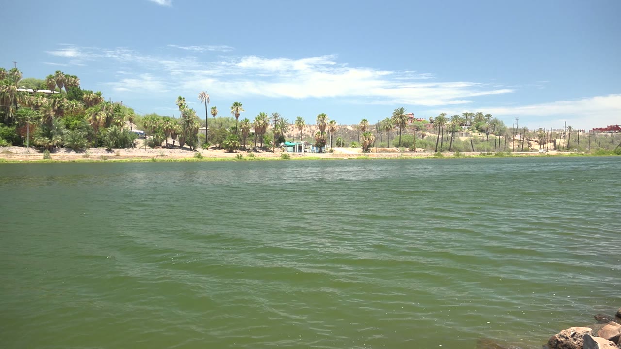 By 0:02 the Mulege waterfront shows more open water in front of palm trees, sparse shoreline buildings, pale hills, and no visible people.