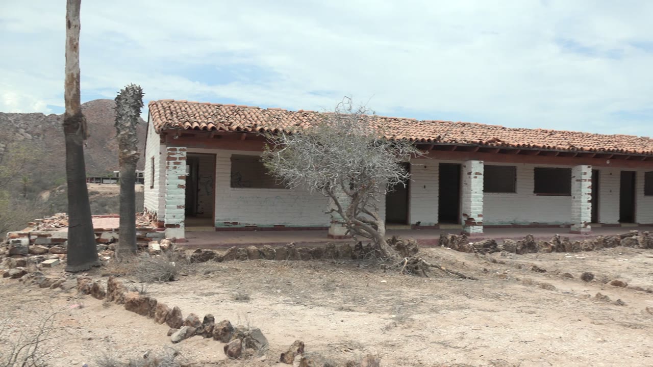 Opening frame of a white desert house in Mulege, Baja California Sur, with a red tiled roof, sandy rock-edged yard, dry trees, distant hills, and no visible people.