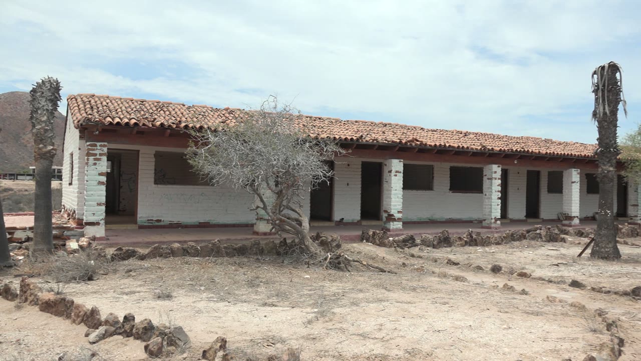 Rightward pan view of the Mulege desert house with a long white wall, red tile roof, dark openings, a dry tree near the porch, a palm trunk at right, and no visible people.
