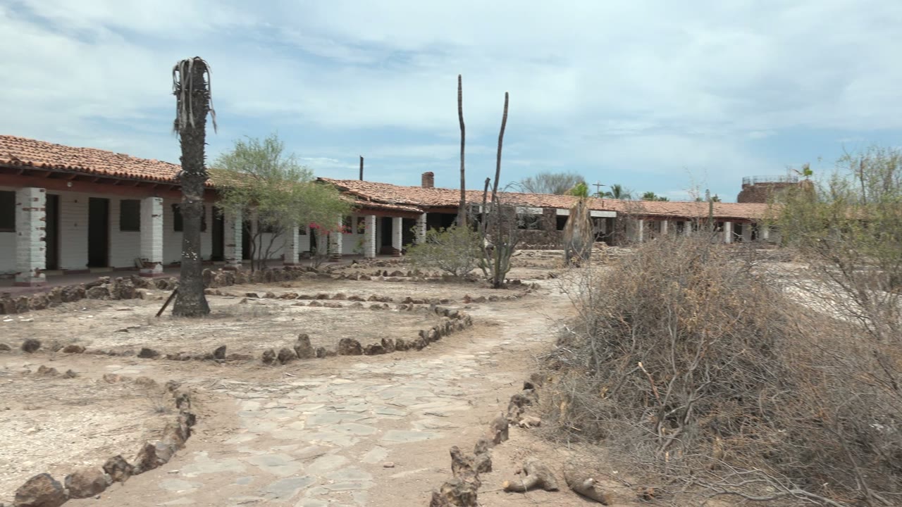 Courtyard path and dry brush in front of red-roofed adobe buildings.