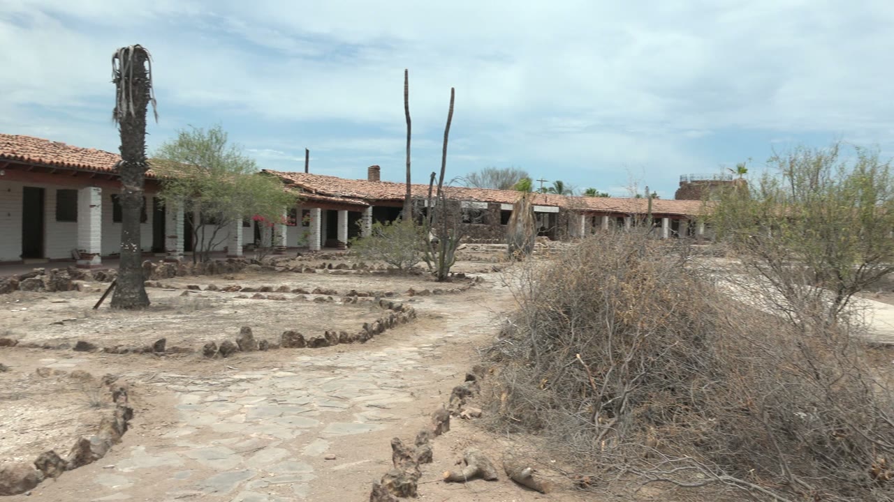 The pan continues across the Mulege courtyard with the adobe building row stretching behind two tall cactus trunks, desert shrubs, low stone borders, and no visible people.