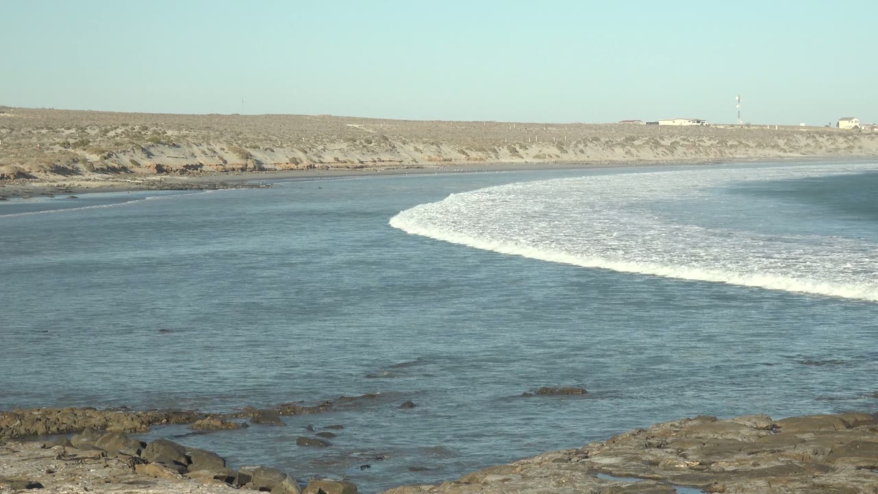 A curved white wave breaks across calm blue water at La Bocana, Baja California Sur, with rocky foreground, low arid coastline, pale sky, and no visible people.
