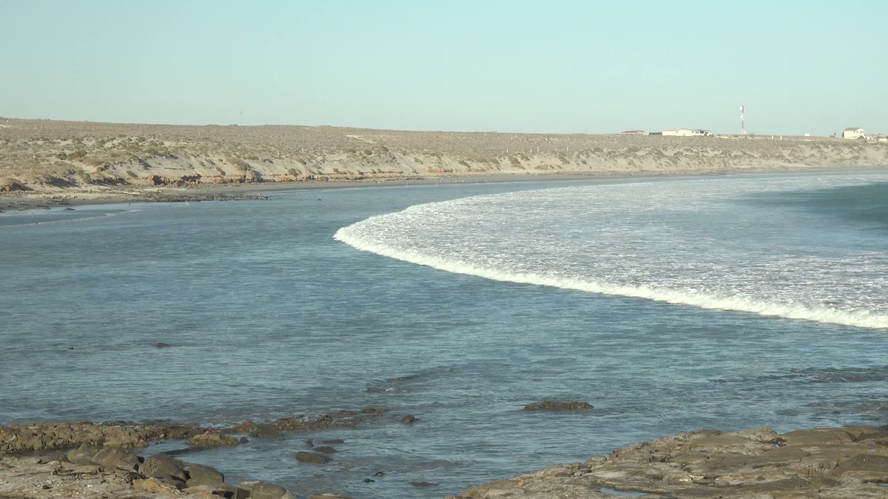 A breaking wave arcs through the midground water near La Bocana, with scattered rocks visible through the surface, a flat arid coastline behind it, and no visible people.