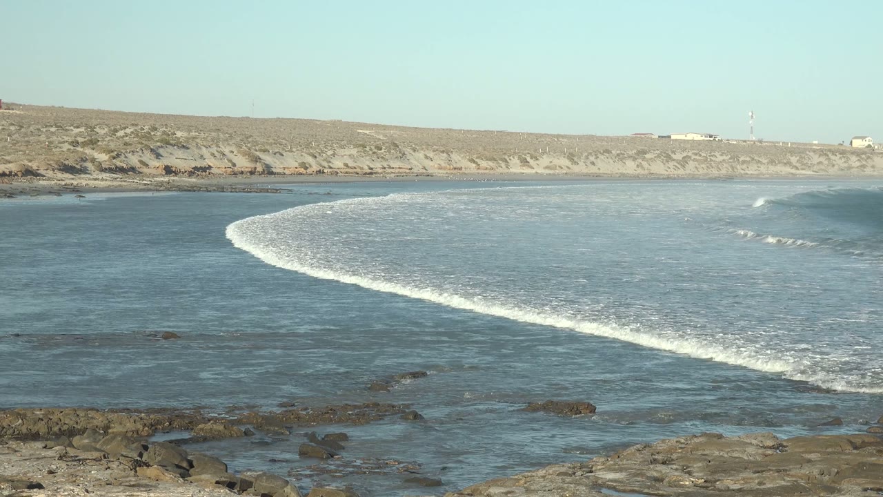 At 0:00 in La Bocana, Baja California Sur, a white wave line curves across blue water toward dark foreground rocks, with a sandy cliff and no visible people.
