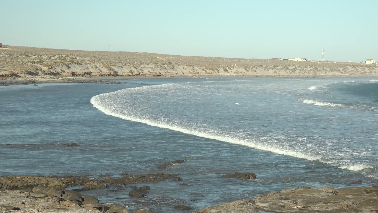 White surf line stretching across blue water near foreground rocks.