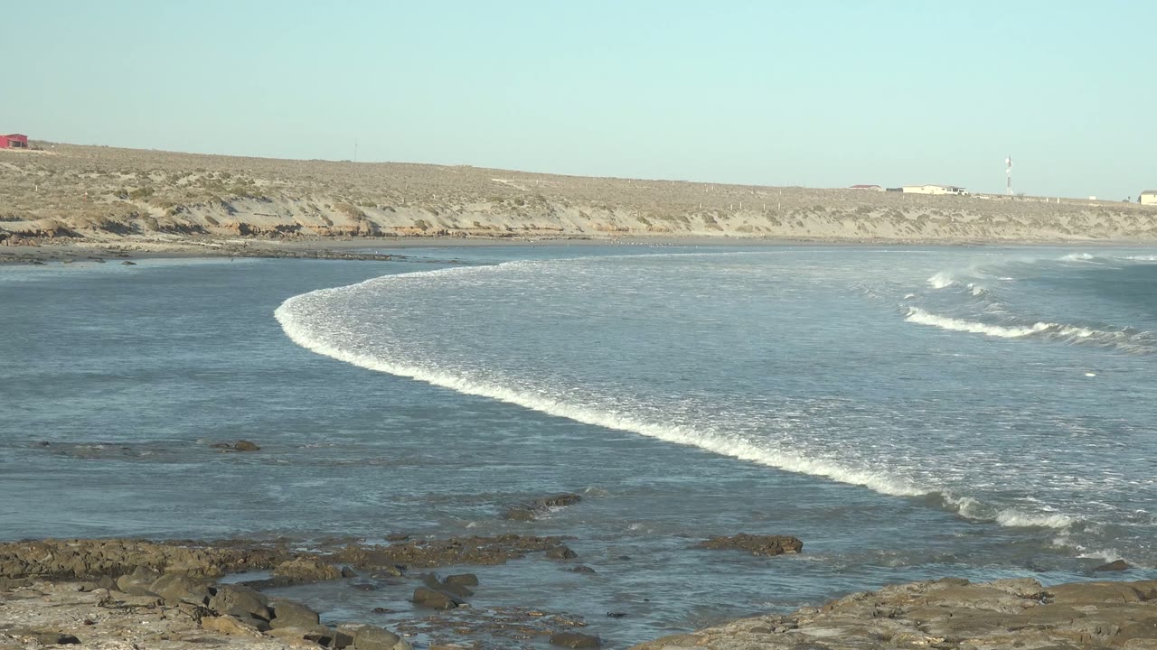 At 0:02 in La Bocana, Baja California Sur, broken whitewater appears to the right while the curved wave front continues toward the rocky foreground, with no visible people.