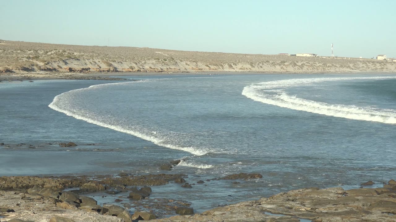 At 0:00 in La Bocana, Baja California Sur, a curved band of white surf moves across blue water toward dark foreground rocks, with no visible people.