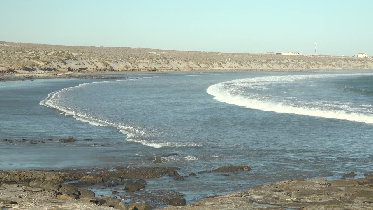 At 0:02 in La Bocana, Baja California Sur, the surf curls across the right side of the bay while shallow water and exposed rocks fill the foreground, with no visible people.