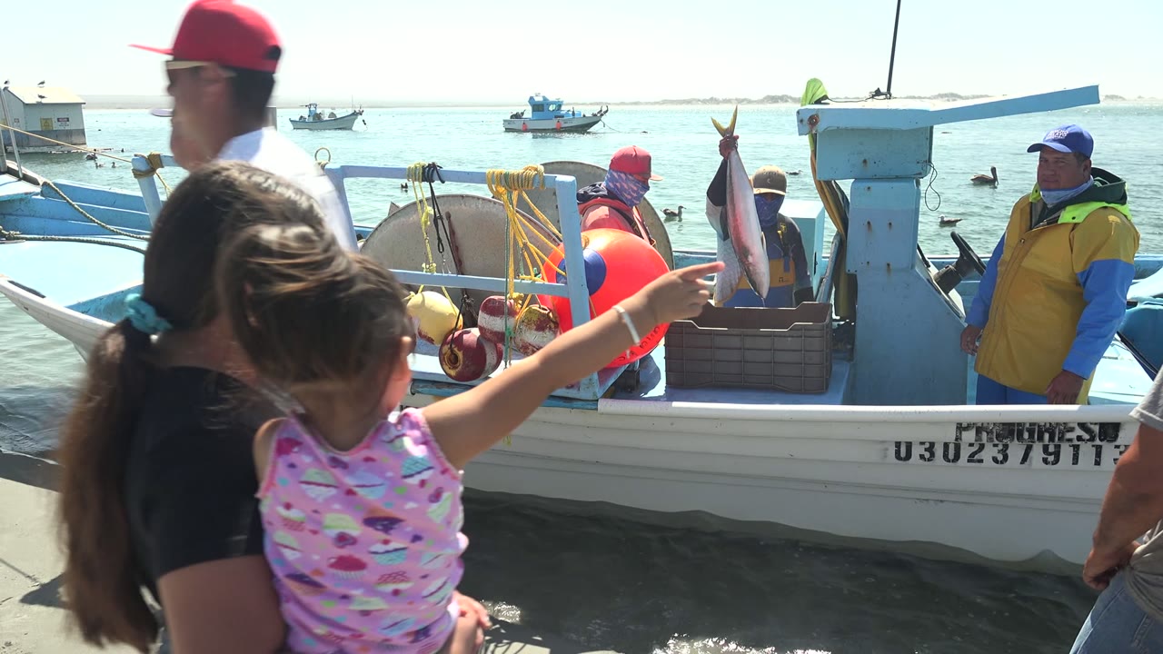 The child points toward a fish being held near the La Bocana fishing boat as adults stand close to the dock and water.