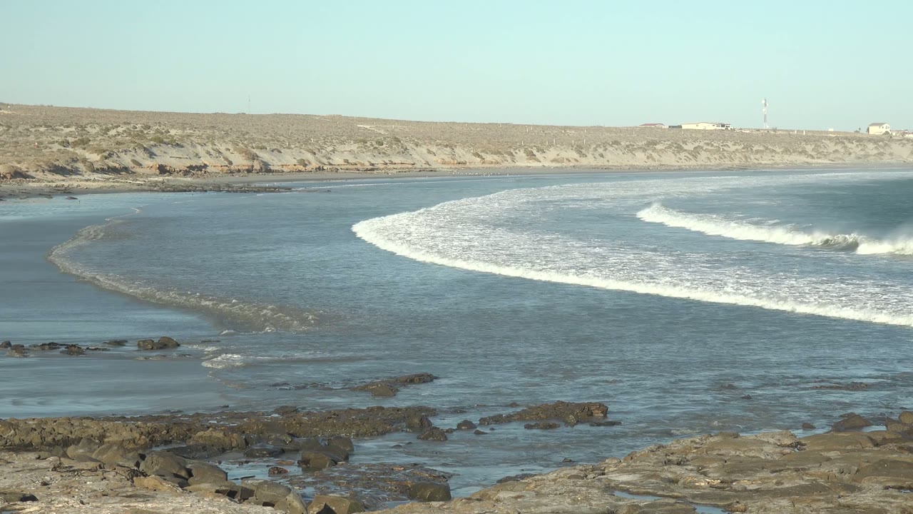 At La Bocana, Baja California Sur, a wide view shows white surf curling across blue water toward foreground rocks, with a sandy cliff on the horizon and no visible people.