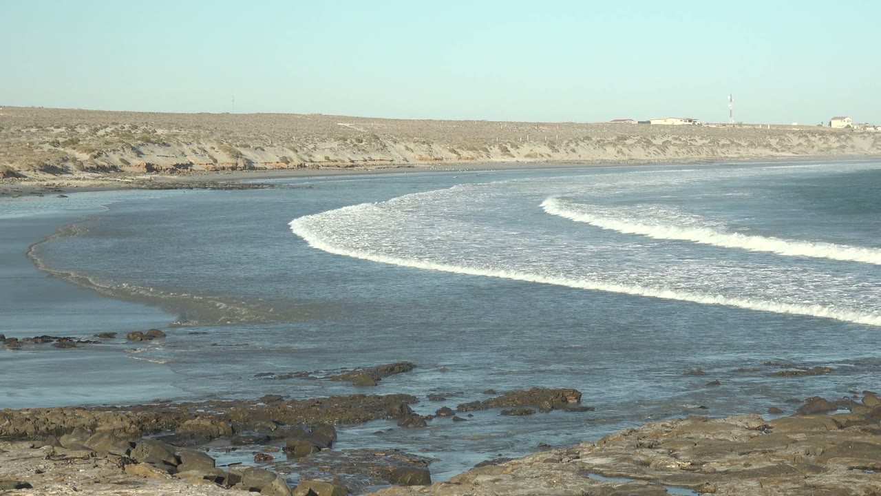 At La Bocana, Baja California Sur, two white wave bands sweep diagonally through the blue water toward foreground rocks, with the sandy cliff and sparse vegetation across the background and no visible people.