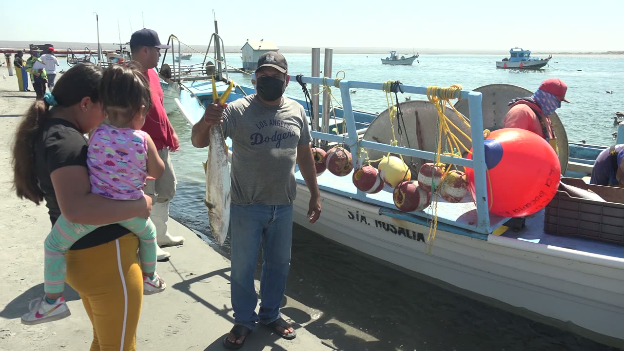 Woman holding a child near a fisherman and a boat with bright buoys.