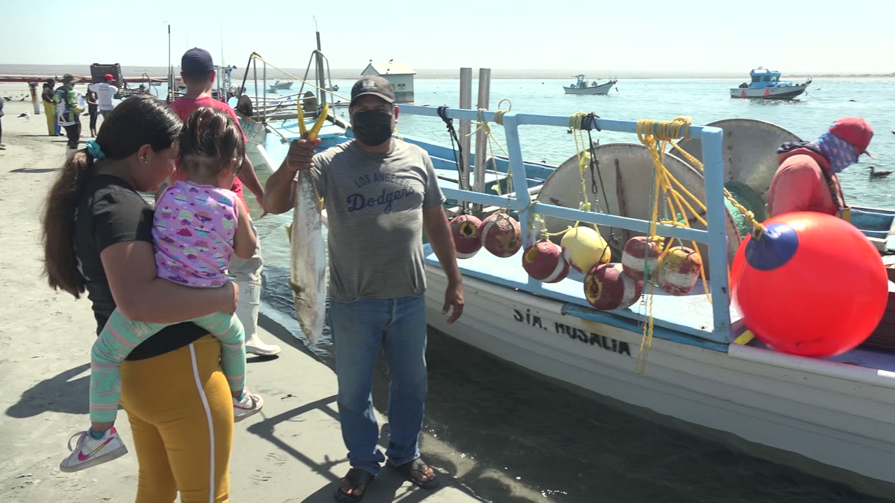 At La Bocana harbor, the fisherman remains beside the boat with the fish while the woman and child face toward him and the boat's blue rail, red buoy, and hanging gear fill the right side.