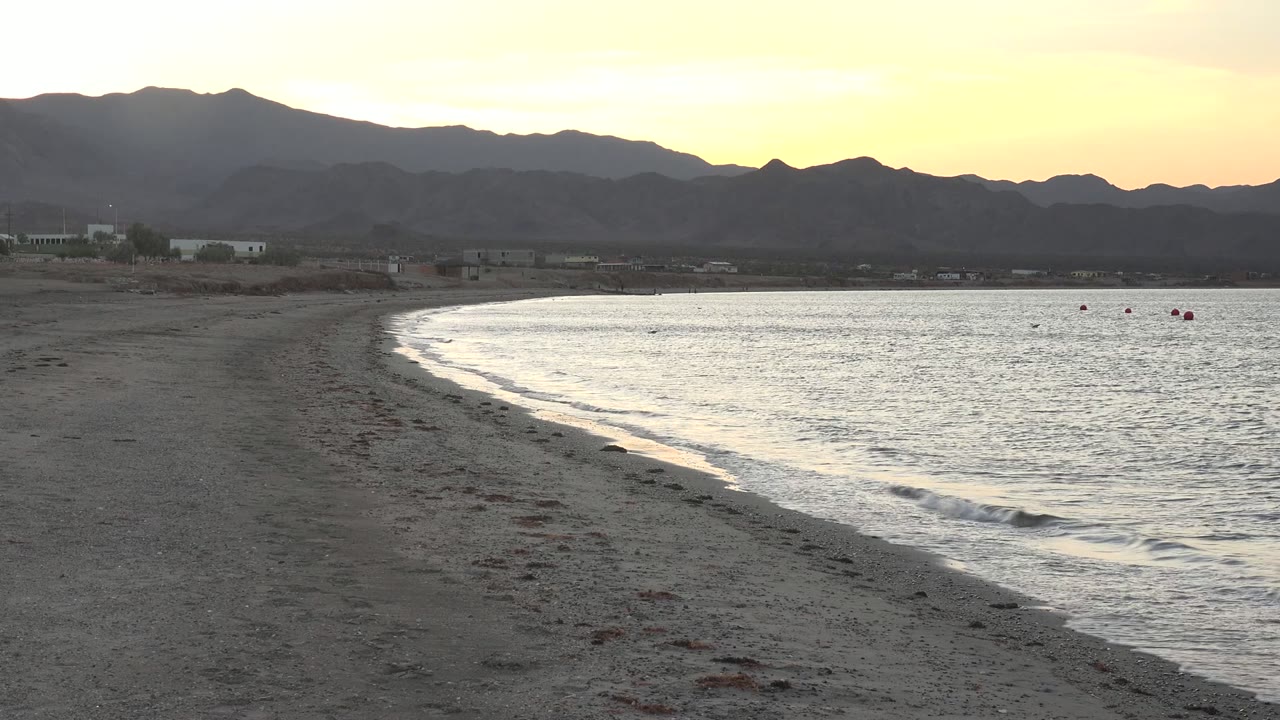 Sandy shoreline and small waves below a pale sunset sky