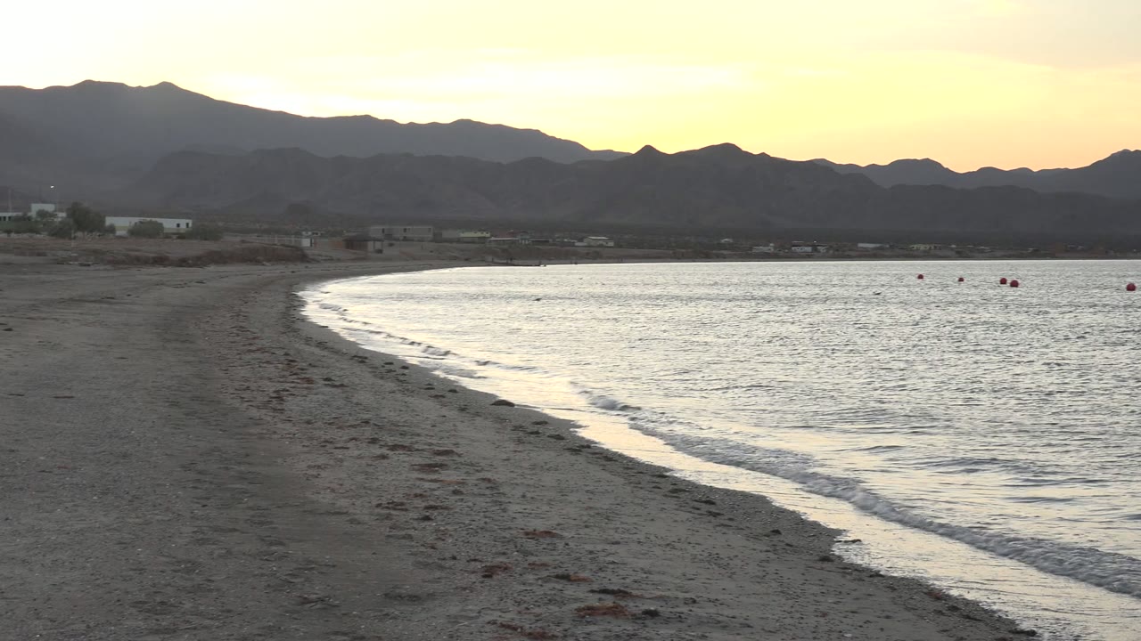 Bahia De Los Angeles beach view with the wave edge curving into calm water, several small buoys offshore, dark mountains behind the bay, and no visible people.