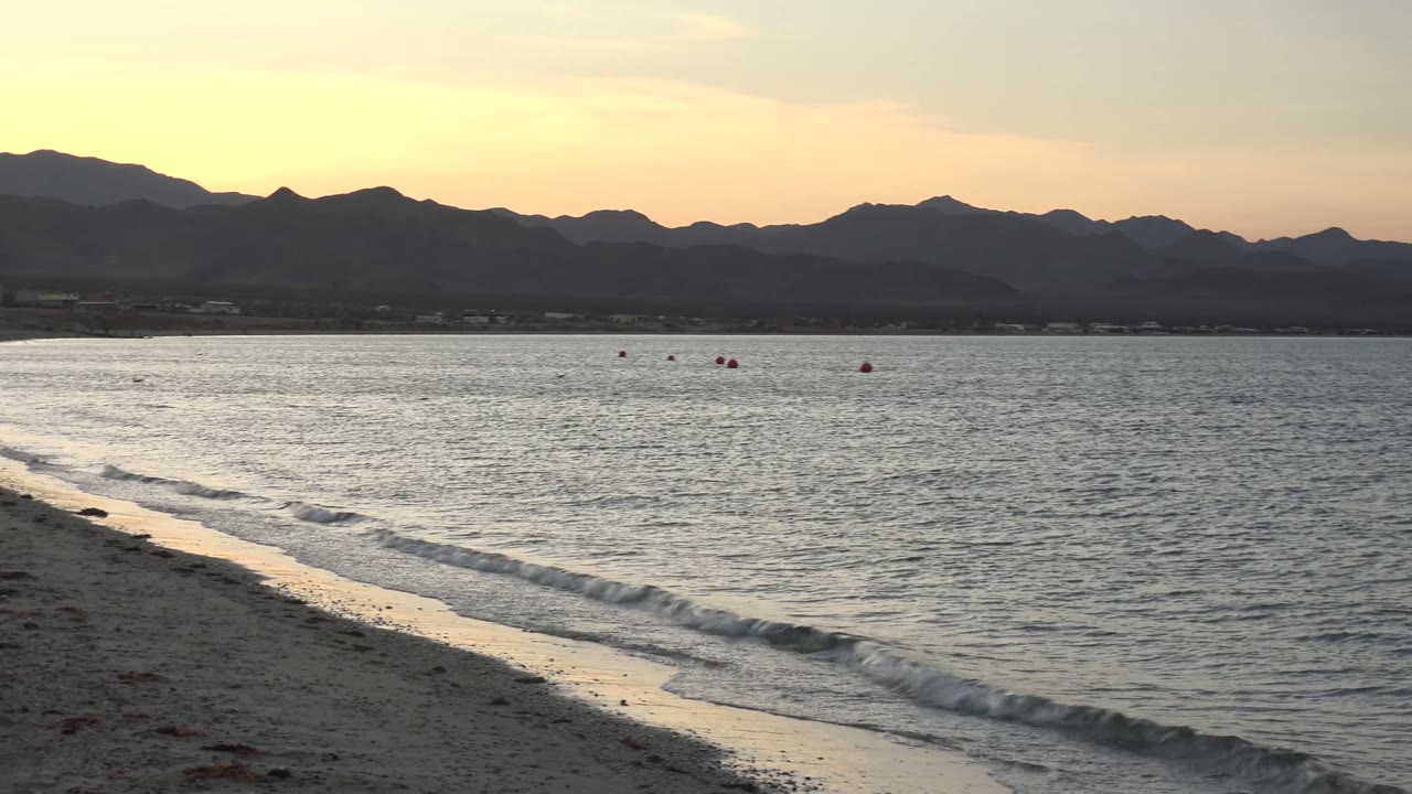 Bahia De Los Angeles beach at sunset with a diagonal sandy shoreline, small waves, red buoys offshore, distant mountains, and no visible people.