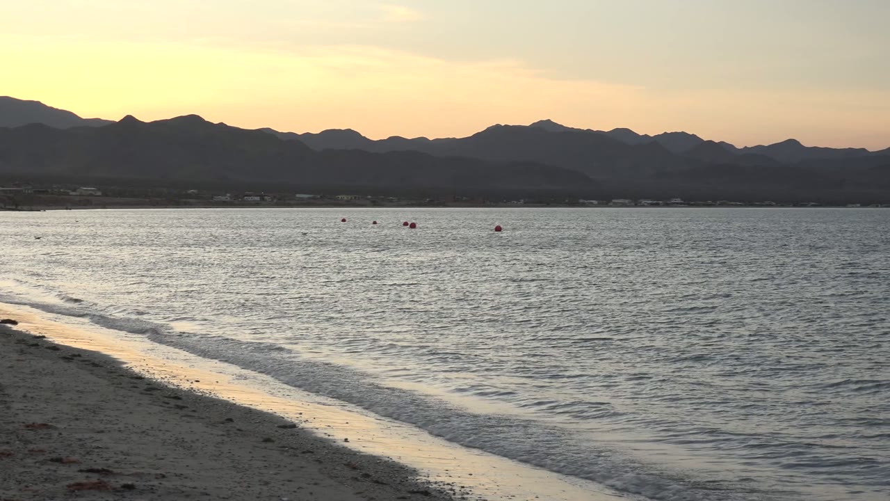 Bahia De Los Angeles shoreline at 0:01 with a brighter strip of reflected sunset light along the wet sand, calm water, red buoys, mountains, and no visible people.