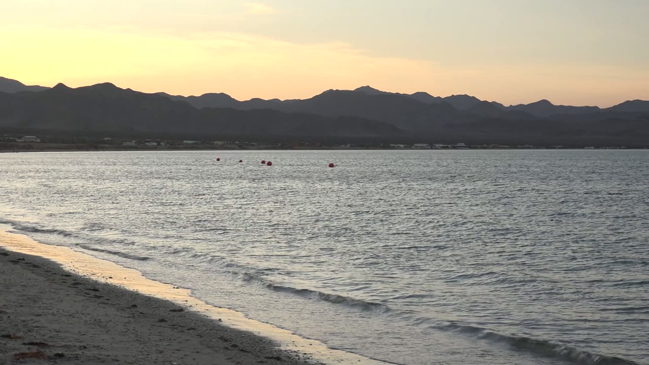 Bahia De Los Angeles water and beach at sunset with the surf line slightly farther right, scattered red buoys offshore, layered mountains, and no visible people.