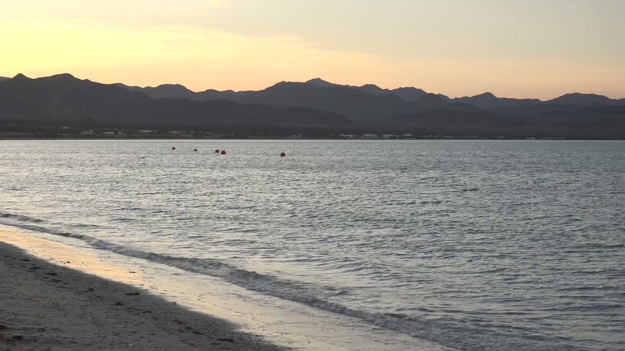 Sandy beach and rippled ocean under a yellow sunset sky with mountain silhouettes.