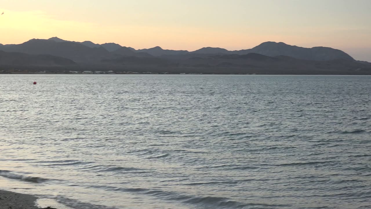 Bahia De Los Angeles shoreline at dusk with low waves across the foreground, open water through the center, mountain silhouettes on the horizon, and no visible people.
