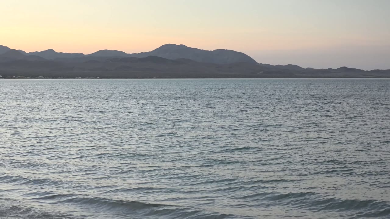 Bahia De Los Angeles ocean water fills the foreground with small ripples, distant mountains sit in silhouette under a pale dusk sky, and no visible people appear.