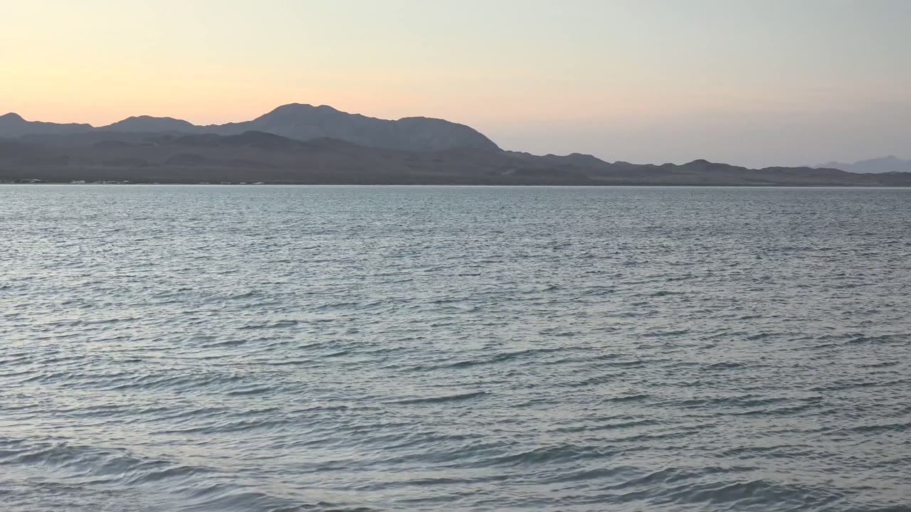 At two seconds, low ocean waves cross the foreground while the Bahia De Los Angeles mountain ridge remains dark beneath a peach-toned dusk band, with no visible people.