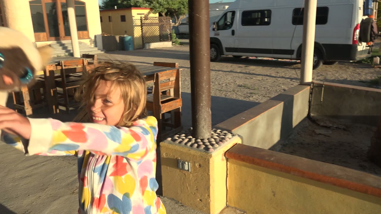A smiling child holds the small rabbit forward toward the camera, with patio furniture, sandy ground, a palm trunk, and a parked van visible behind in San Carlos, Mexico.