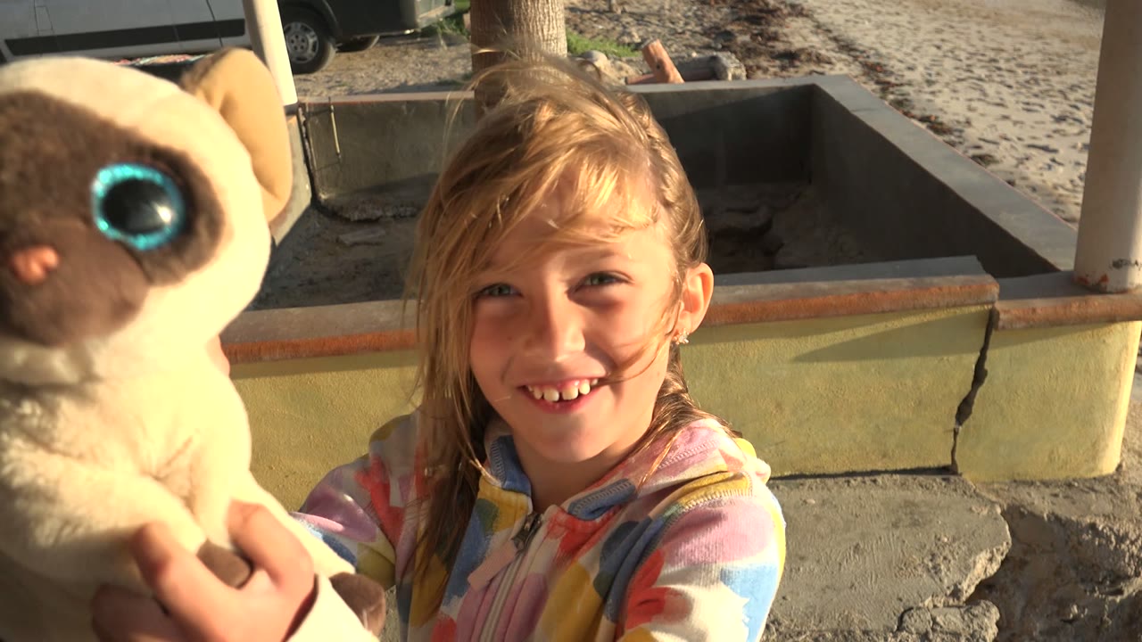 A young girl in a colorful jacket smiles in San Carlos, Mexico while holding a plush animal close to the camera beside a concrete patio and sand.
