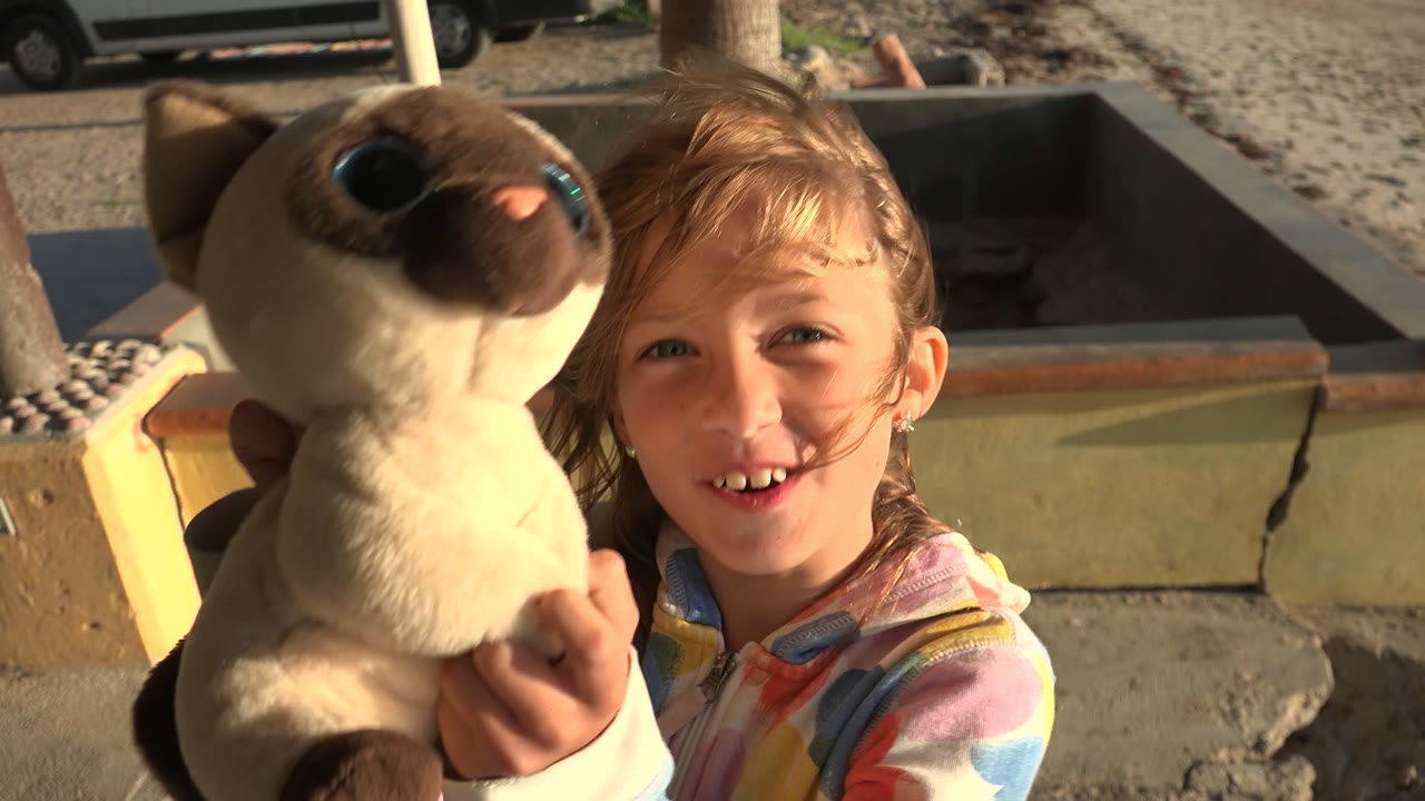 Girl holding a stuffed animal toward the camera on a sunny patio.