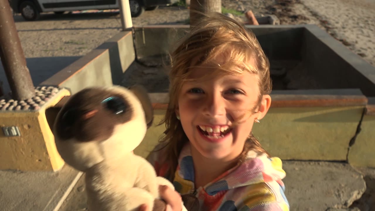 The girl smiles in San Carlos, Mexico while the plush toy blurs at the edge of frame and the patio wall, sand, and outdoor seating stay behind her.