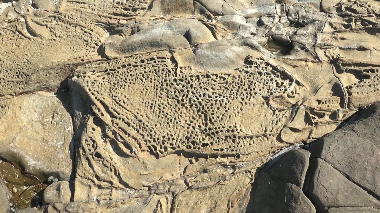 The pitted La Bocana rock remains centered, with clustered holes, chipped edges, and side shadows around the surrounding stones, with no visible people.