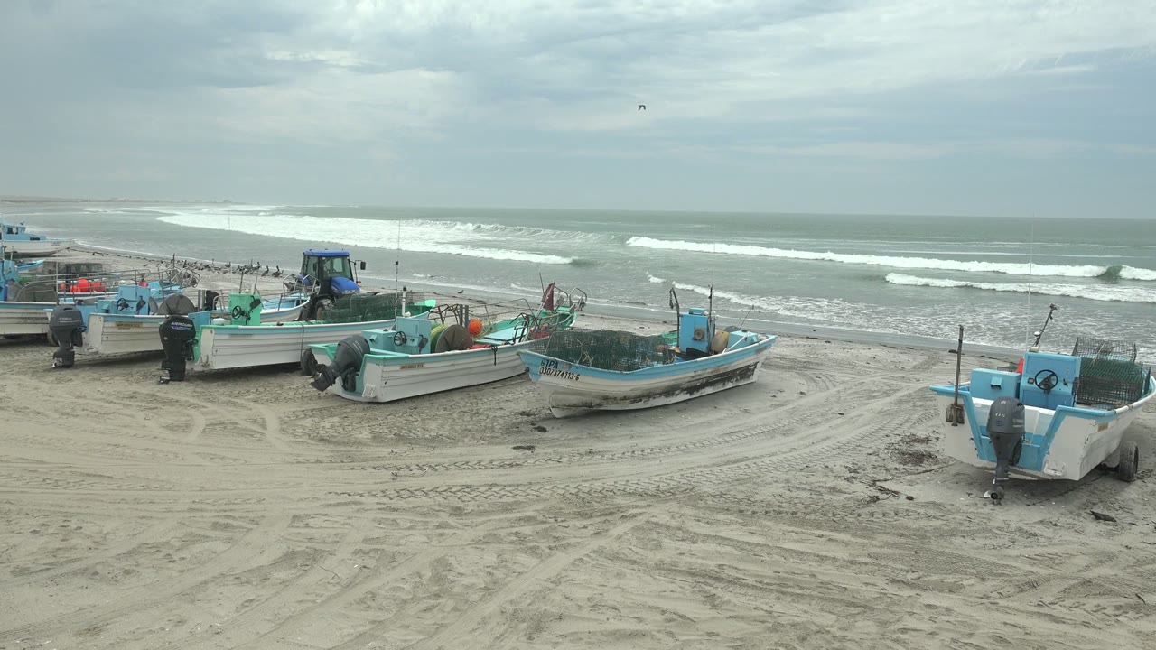 Blue and white fishing boats rest on tire-marked sand at Punta Abreojos, Baja California Sur, with breaking waves and an overcast sky behind them, no visible people.