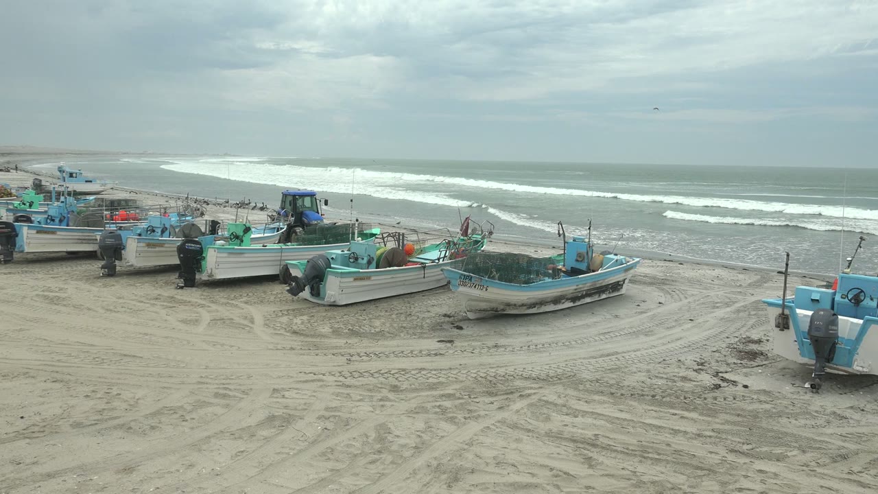 Blue-trimmed fishing boats line the sandy beach at Punta Abreojos while white wave breaks stretch across the gray water, with no visible people.