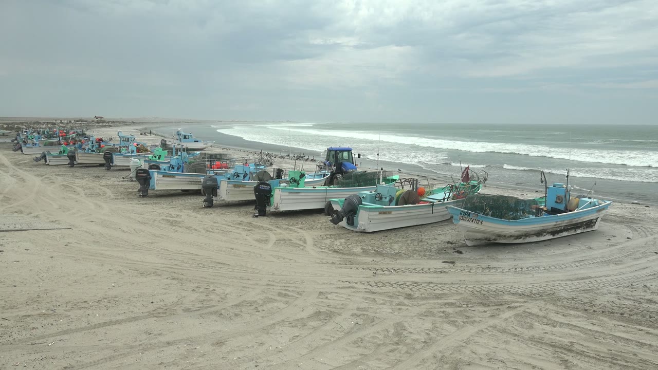 Wide view of fishing boats on the sandy shore at Punta Abreojos, Baja California Sur, with surf behind them, tire tracks in the foreground, and no visible people.