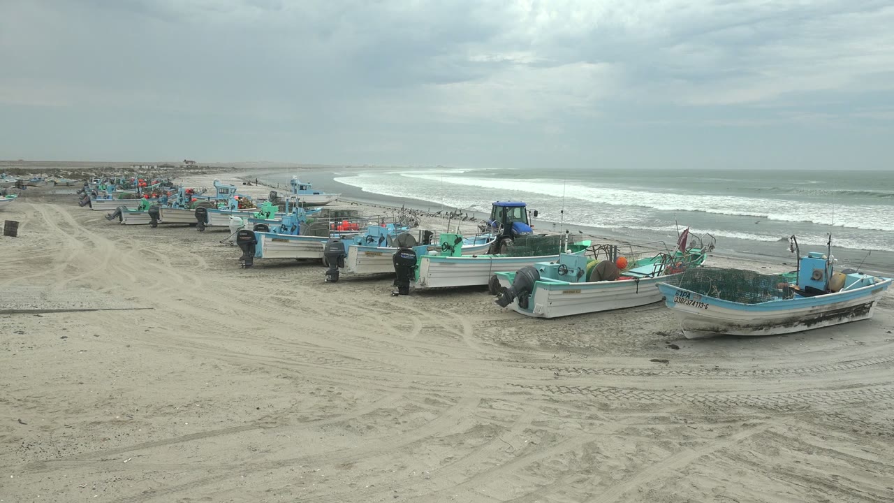 Fishing boats remain lined along the Punta Abreojos sandy shore, with tire-marked sand in front, a low ocean horizon behind, and no visible people.