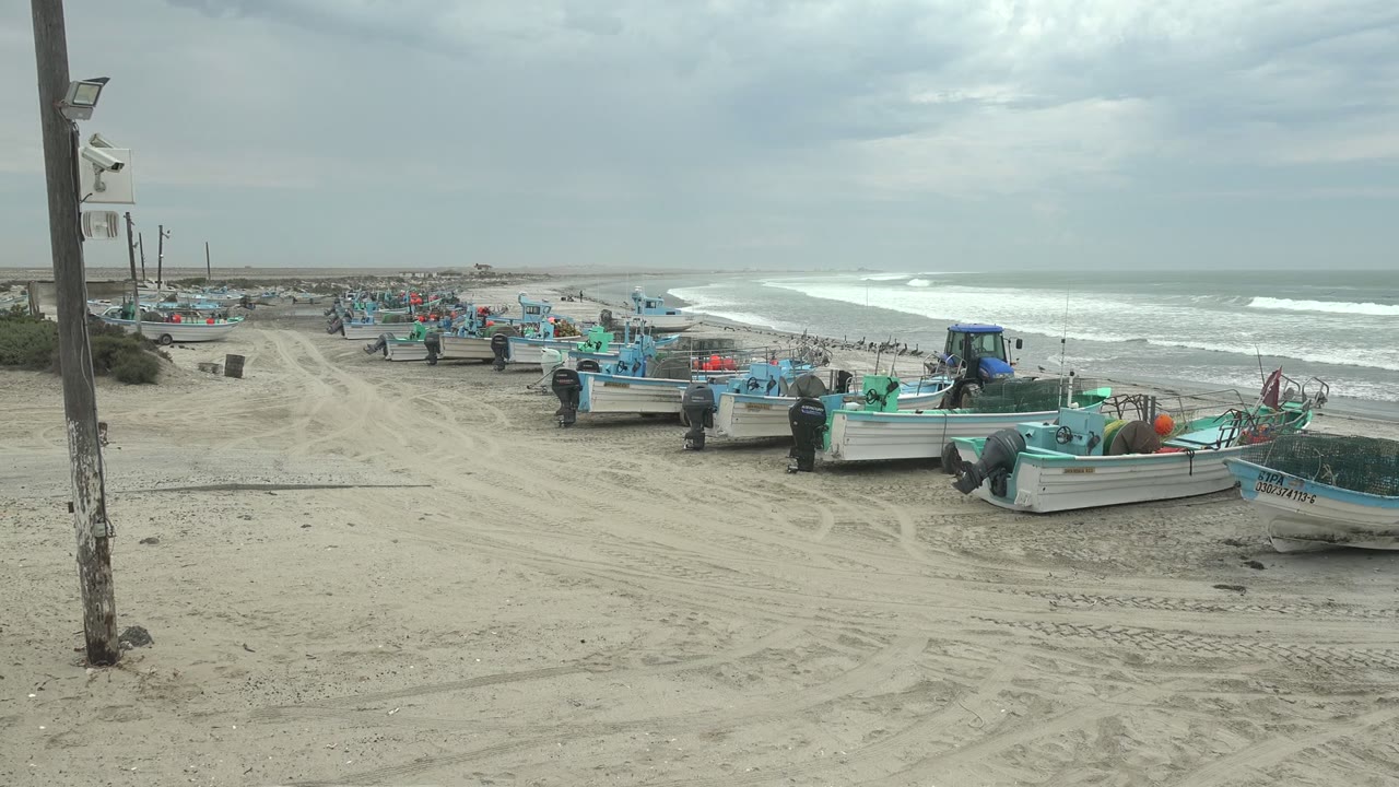 At Punta Abreojos, Baja California Sur, fishing boats line the sandy shore with people near the boats, a utility pole at left, and white surf rolling in under an overcast sky.