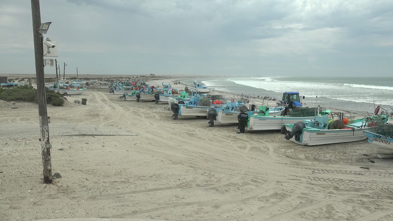At Punta Abreojos, Baja California Sur, the frame shows parked fishing boats stretching down the beach, a few people near the hulls, surf breaking to the right, and muted gray-blue overcast light.
