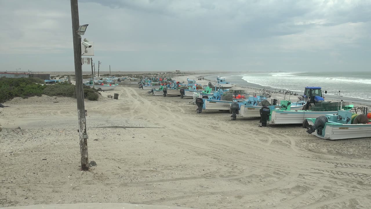 Wide view of fishing boats on a sandy beach in Punta Abreojos, Baja California Sur, with a foreground utility pole, people near the boats, and surf rolling in the background.