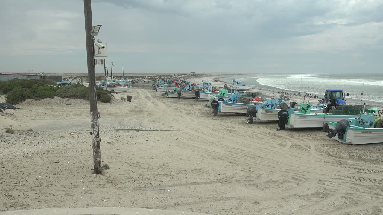 Parked blue and white fishing boats line a sandy shore near ocean waves.