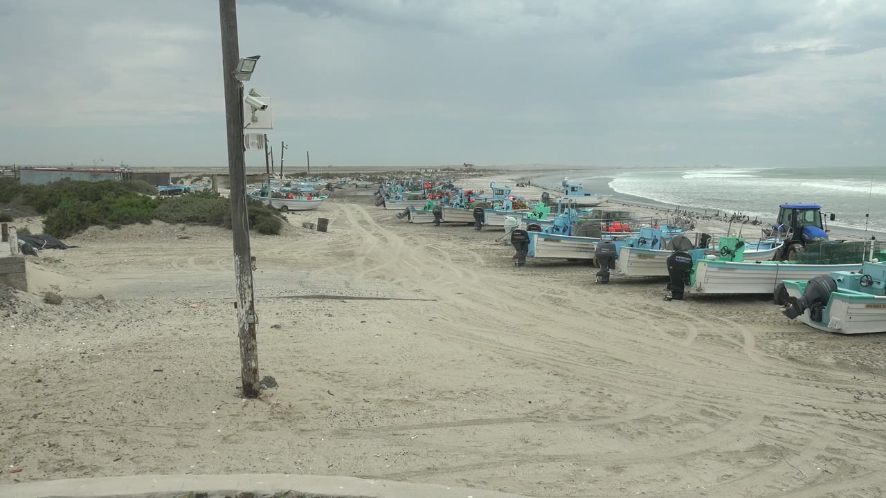 Wide Punta Abreojos shore frame with small fishing boats in the midground, a person near a boat at right, and bright surf visible beneath overcast clouds.