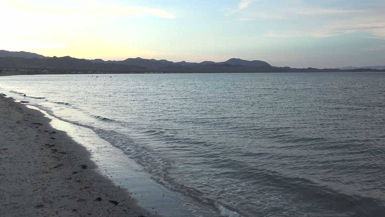 At 0:00, a small wave curls toward wet sand in Bahia De Los Angeles with blue-gray water, a dark mountain horizon, pale sunset sky, and no visible people.