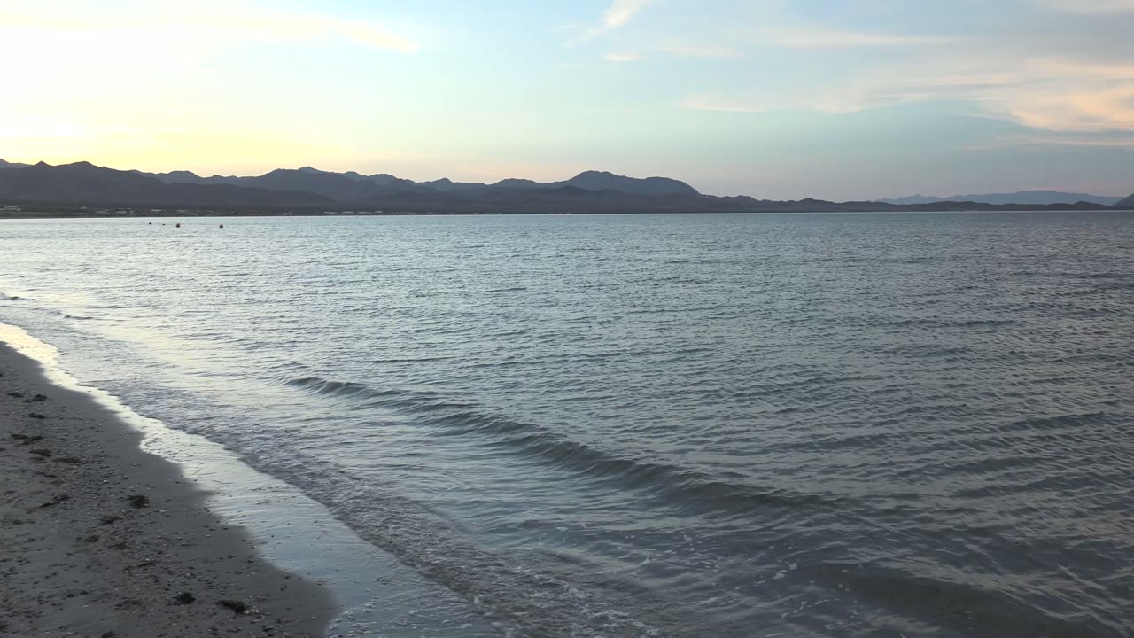 At 0:02, a low wave approaches the sandy edge in Bahia De Los Angeles while open water fills most of the frame beneath distant mountains and no visible people.