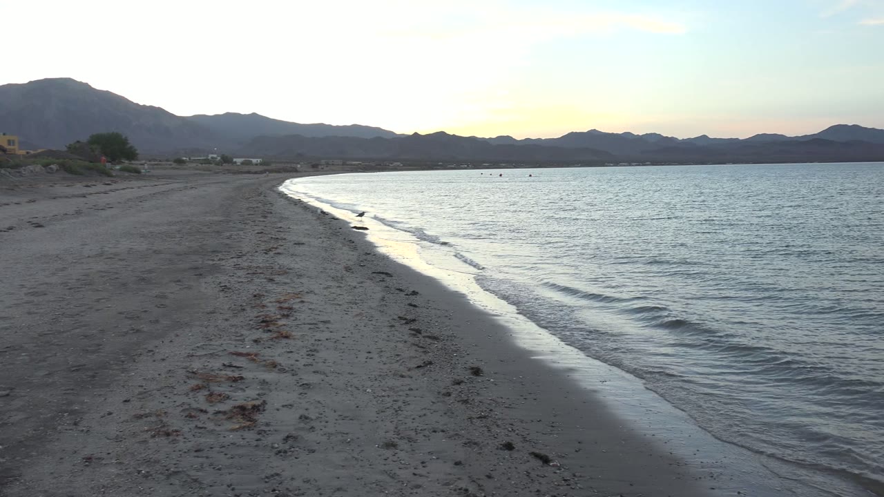 Gentle wave line running along a sandy shore with mountains beyond the water.