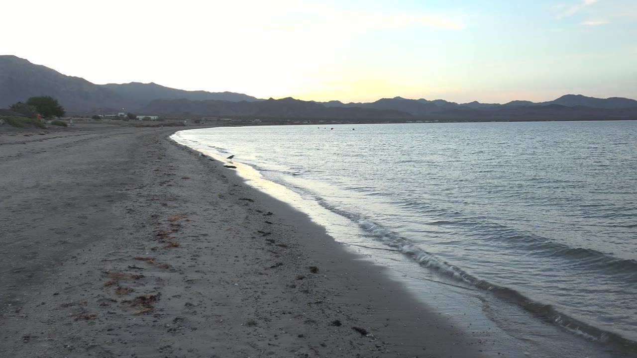 At 0:02, foam curls onto the sandy beach in Bahia De Los Angeles while calm water stretches toward silhouetted mountains and no visible people appear.