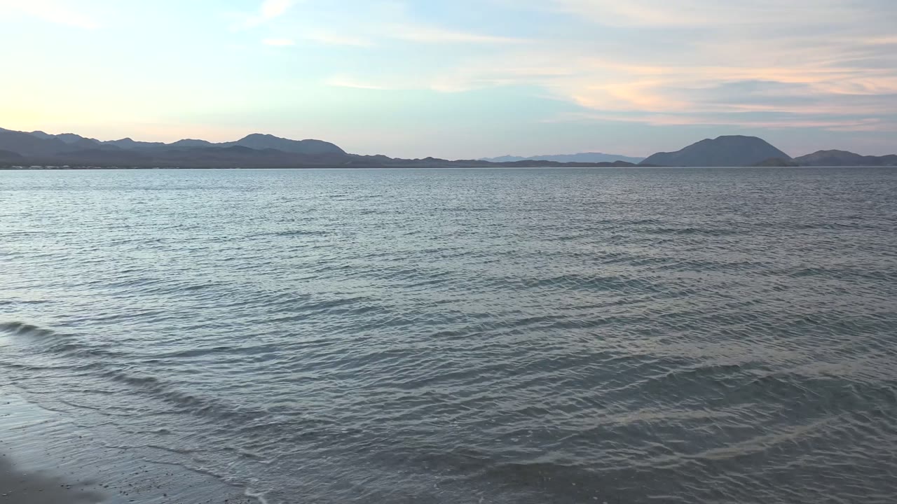 Gentle ocean waves fill the foreground at Bahia De Los Angeles, with low islands on the horizon under a pale partly cloudy sky and no visible people.