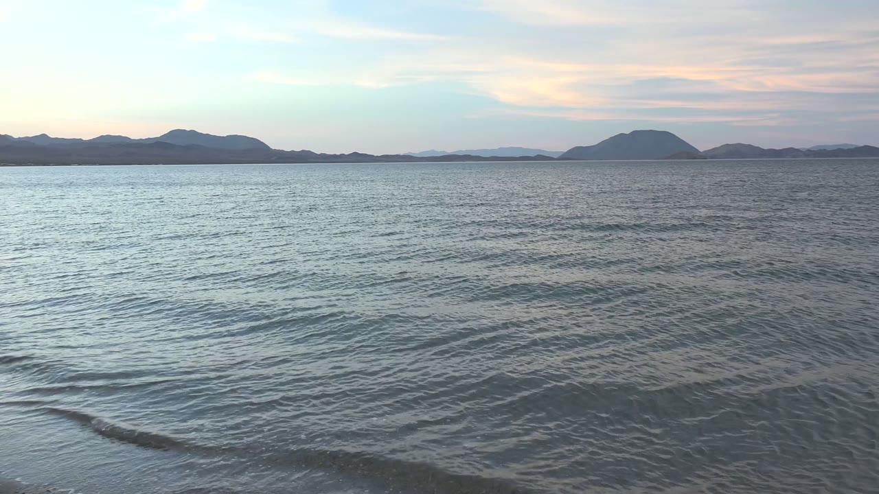 A thin wave line forms across the right foreground at Bahia De Los Angeles, with calm open water leading to distant hills and no visible people.