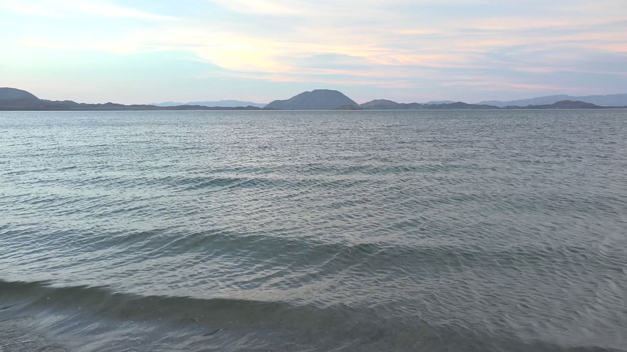 Gentle ocean waves fill the foreground at Bahia De Los Angeles, with distant island silhouettes under a pale pastel sky and no visible people.